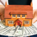 A person holding a model house on top of money.