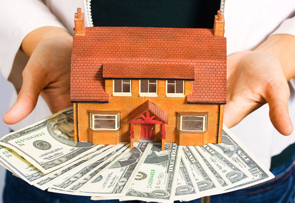 A person holding a model house on top of money.